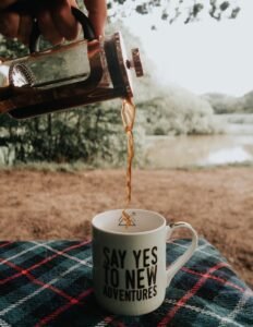 Coffee poured into a mug with adventure-themed text on a plaid blanket outdoors.