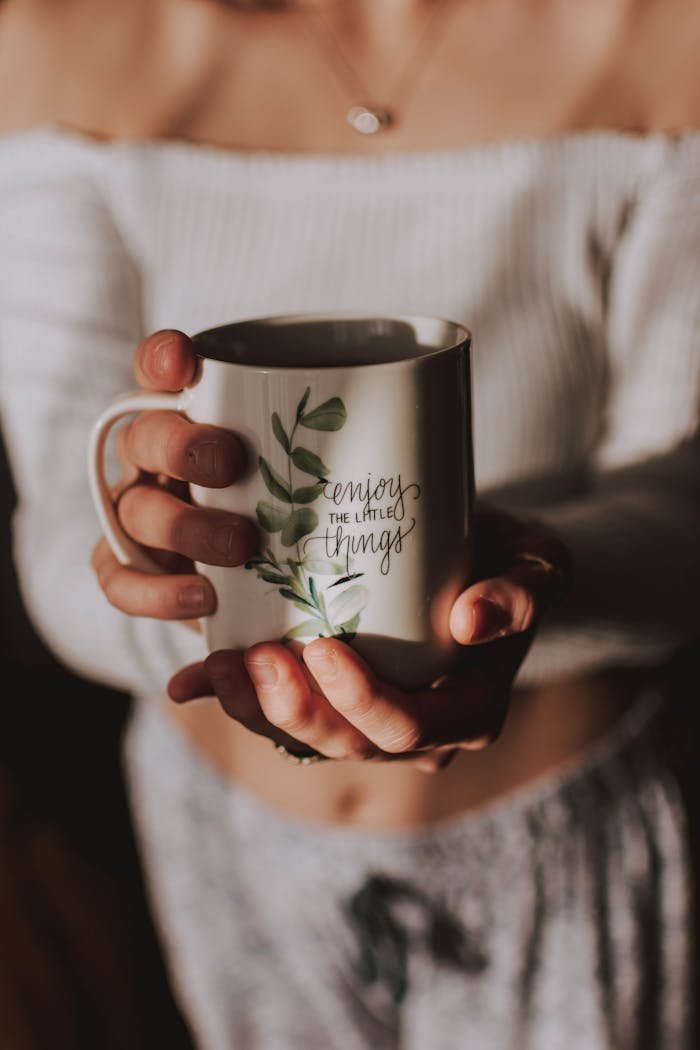 A woman holds a mug with 'enjoy the little things' in a cozy, warm setting.